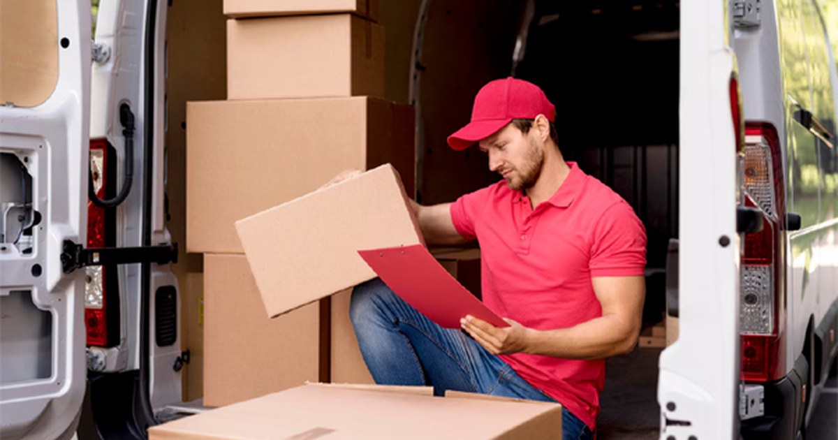 Repartidor con gorra y polo rojos revisa el albarán junto a las cajas apiladas en la furgoneta de reparto de mercancías