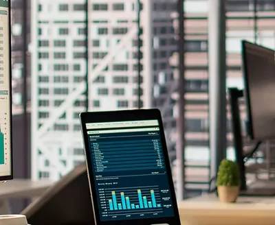 Oficina con un monitor y una tableta que muestran gráficos y cuadros de mando, con edificios de la ciudad al fondo