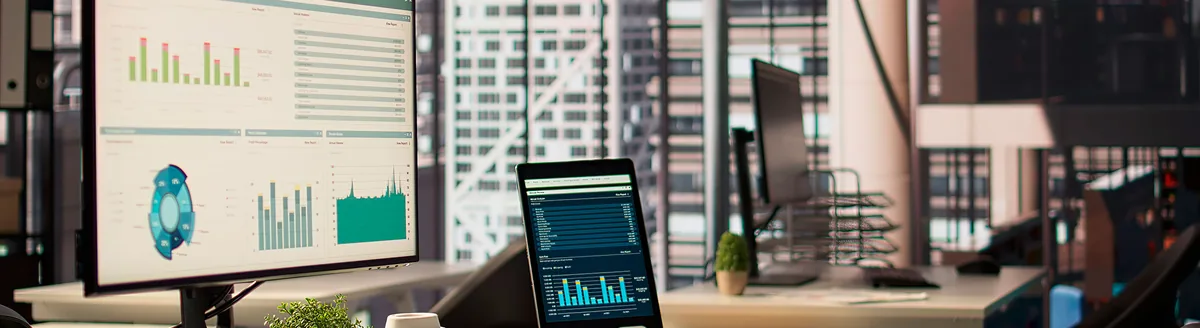 Oficina con un monitor y una tableta que muestran gráficos y cuadros de mando, con edificios de la ciudad al fondo