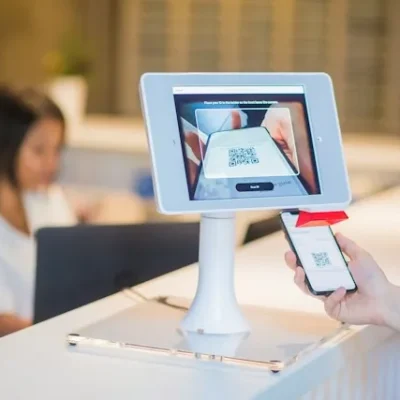 Mostrador de recepción con una tablet en soporte que muestra un código QR y un cliente acercando su móvil, con una recepcionista trabajando al fondo