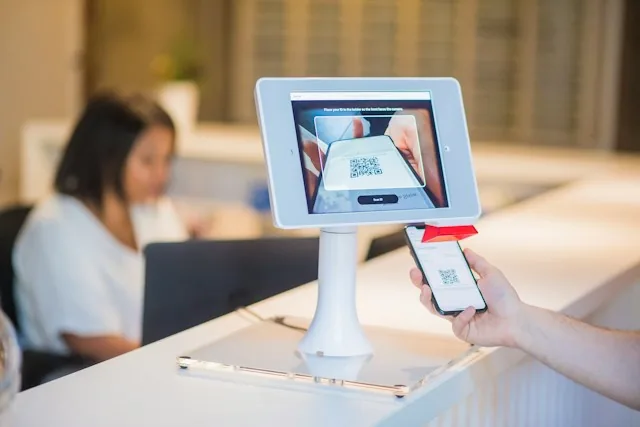 Mostrador de recepción con una tablet en soporte que muestra un código QR y un cliente acercando su móvil, con una recepcionista trabajando al fondo